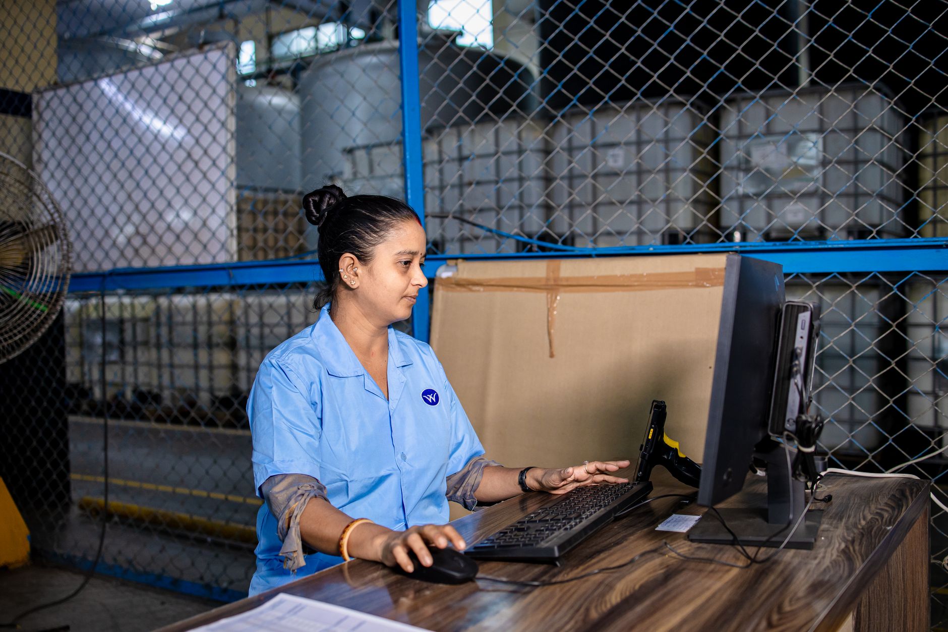 A woman in blue uniform operates a computer inside a factory warehouse, focusing on her ta