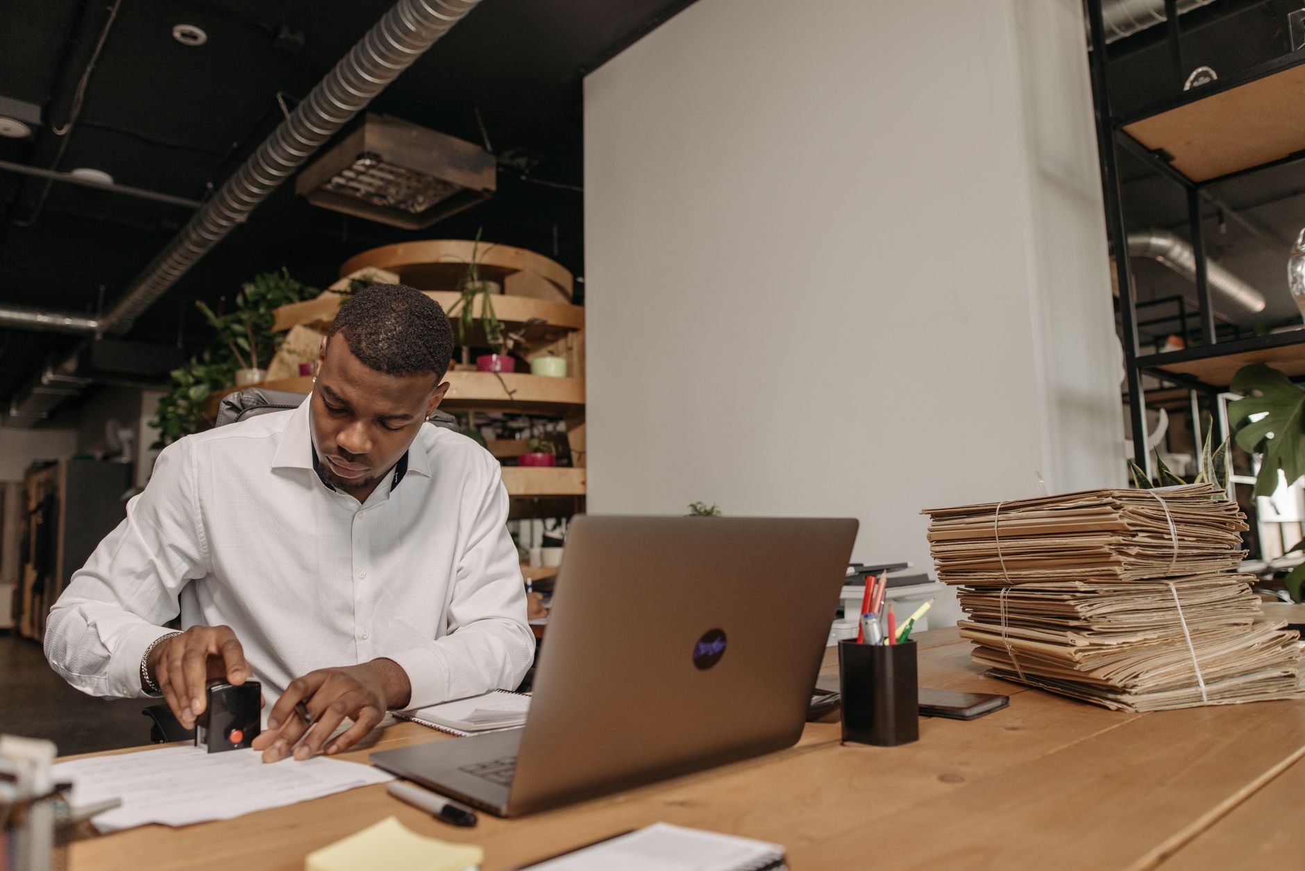 African American man focused on stamping documents at an office desk with a laptop and fol